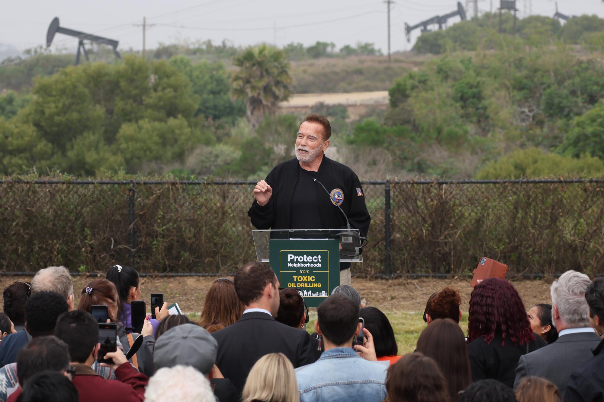 Der ehemalige kalifornische Gouverneur Arnold Schwarzenegger spricht im Freien, während im Hintergrund Ölpumpenheber zu sehen sind.