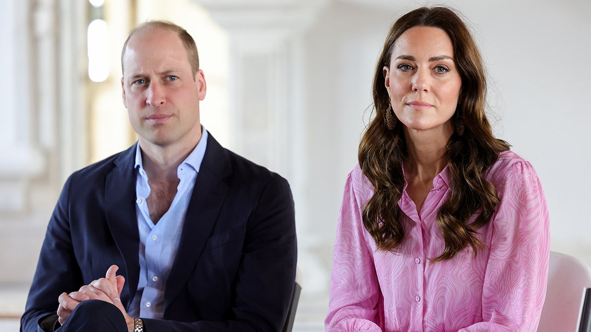 Prinz William im schwarzen Anzug über einem hellblauen Hemd sitzt ernst neben Kate Middleton im rosa Outfit und sieht ebenfalls stoisch aus