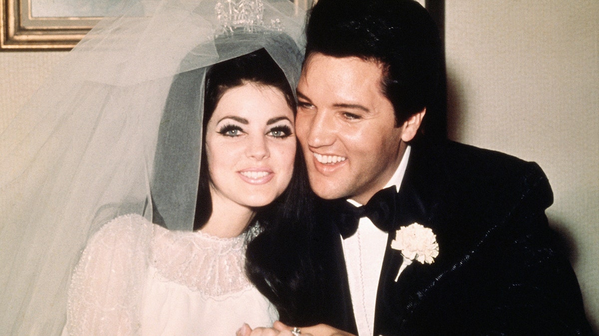 Priscilla Presley im weißen Hochzeitskleid und Tüllschleier lächelt neben Elvis Presley im klassischen Smoking