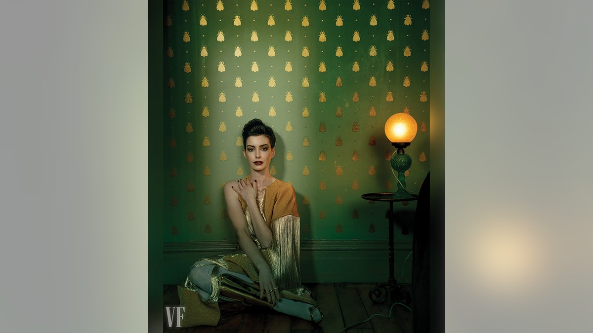 Anne Hathaway in einem gold- und senfgelben Kleid legt ihre Hand auf ihre Schulter und lehnt an einer Wand mit grüner Tapete und goldenen Insekten darauf