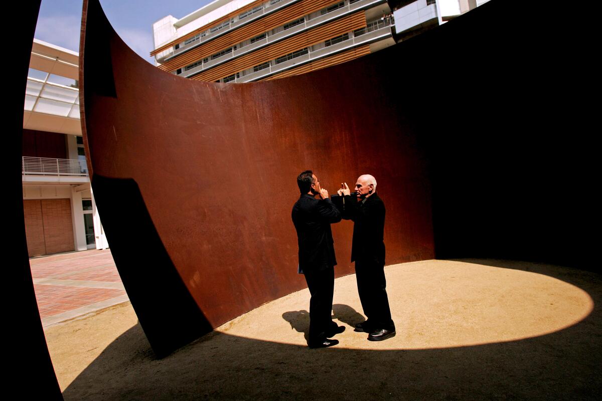 Richard Serra und Franciso Pinto stehen in schwarzen Anzügen in einer massiven gebogenen Stahlskulptur.