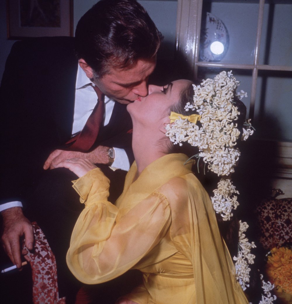 15. März 1964: Die in Großbritannien geborene Elizabeth Taylor sitzt auf einem Sofa und küsst ihren fünften Ehemann, den walisischen Schauspieler Richard Burton, während er sich an ihrem ersten Hochzeitstag über sie beugt.  Taylor trägt ein gelbes Kleid und hat Blumen im Haar.  (Foto von Hulton Archive/Getty Images)