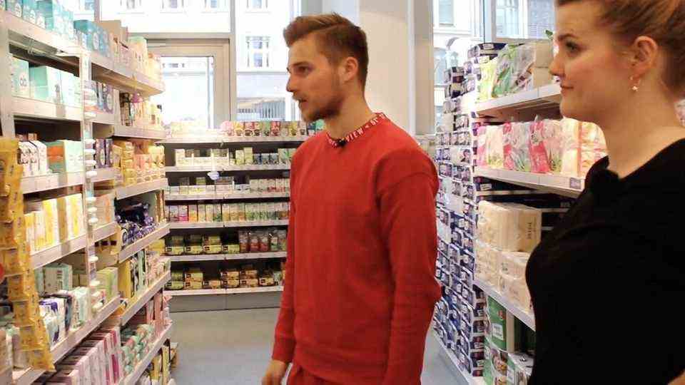 A young man in a red sweat shirt and a young woman in a black top look at a supermarket shelf full of period products