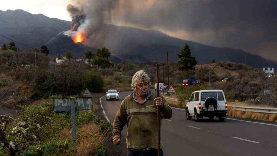 A man stands in front of the active volcano.  Thousands of houses have already been destroyed by the lava.