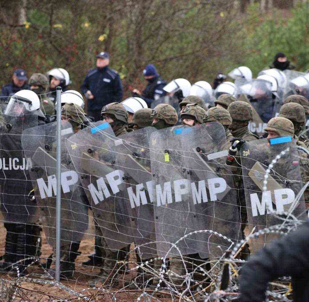 Polish police officers stand behind a barbed wire fence with protective shields while migrants gather at the Belarusian-Polish border