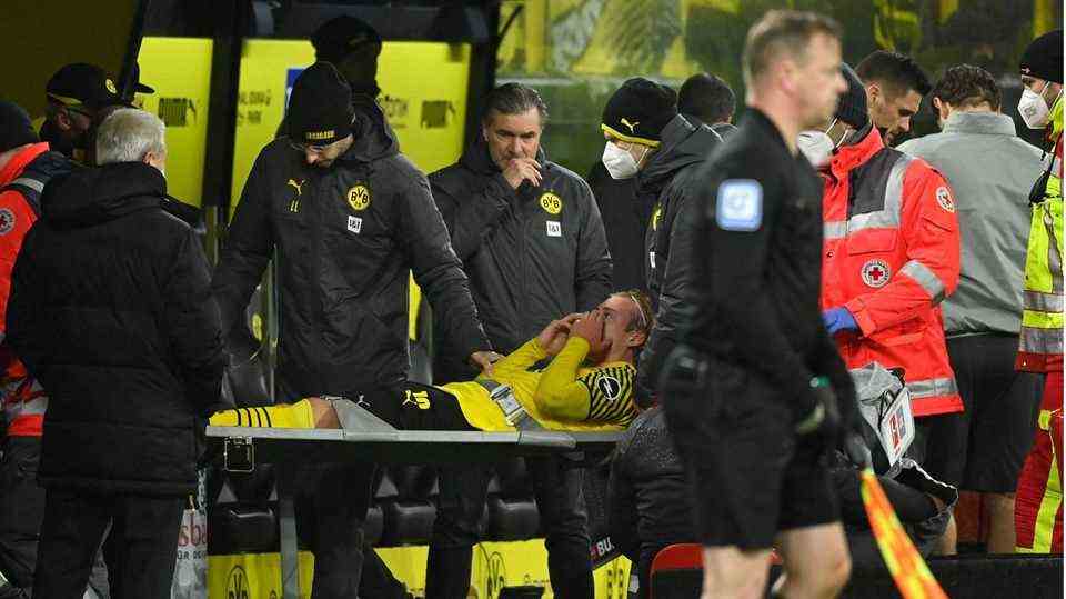 A footballer in a yellow-black jersey and black shorts is being brought from the field on a gray stretcher