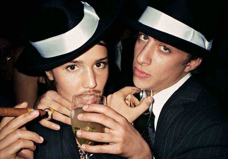 A woman and a man pose in pinstripe suits with hats, wine glasses and cigars