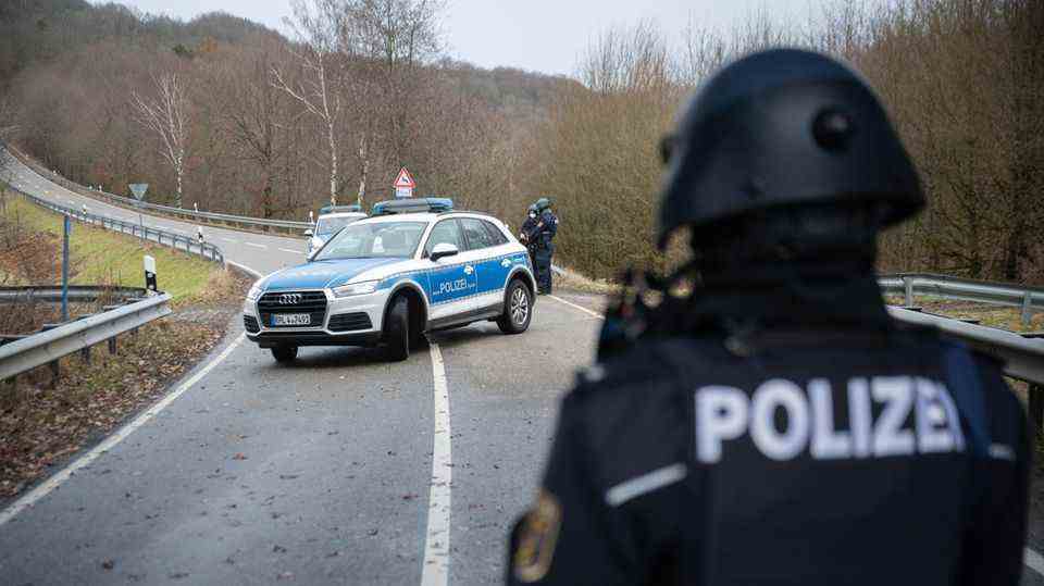 A police officer in riot gear and a helmet looks at a police car parked diagonally across a curving country lane