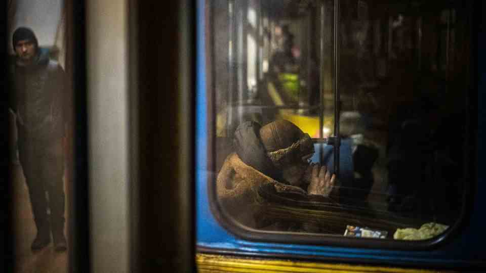 An old woman in a winter coat sits behind a dirty window of a blue and yellow subway train and drinks tea