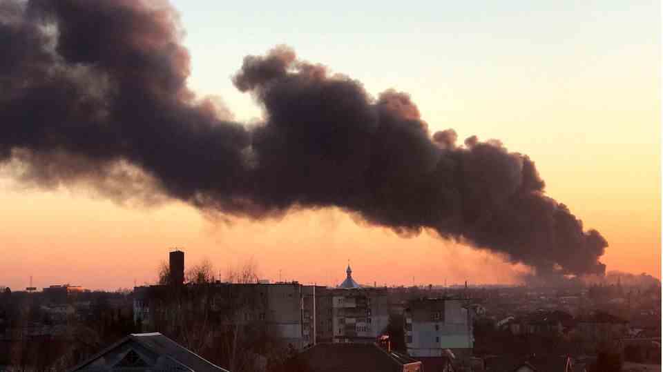 At dawn, a column of black smoke rises from lower right to upper left in the sky above a city