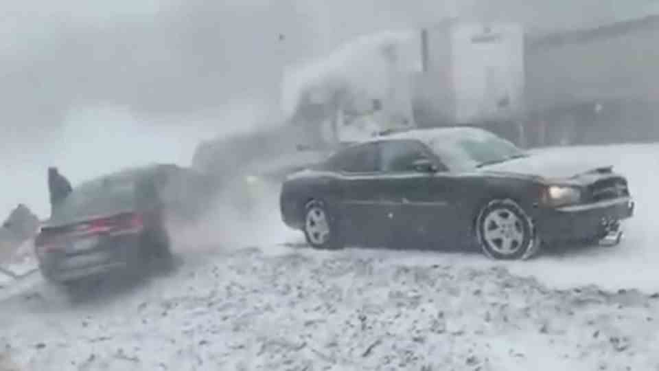 A black sedan is parked on the edge of a snow-covered freeway, behind its rear another car slides backwards past
