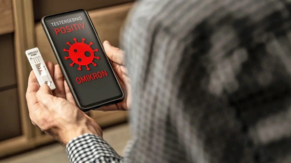 A man is holding a smartphone and a positive rapid coronavirus test.  The black display of the smartphone reads in red: Test result positive Omikron.  © picture alliance / CHROMORANGE |  Michael Bihlmeyer 
