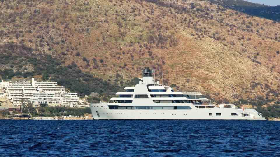 A huge white motor yacht lies in the sun off a sparsely vegetated coast