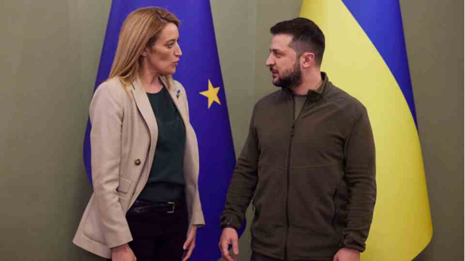 Roberta Metsola and Volodymyr Zelenskyj in front of blue and yellow EU and Ukraine flags