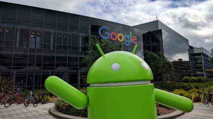 A green android stands in front of Google headquarters (glass building background)