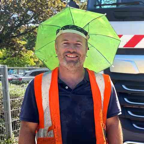 A man wearing a yellow head sunshade that looks like an umbrella that attaches directly to his head stands in front of a truck and smiles at the camera.