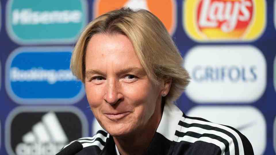 A white woman with a blond parting in the middle and in a black training jacket with white stripes on the sleeves smiles