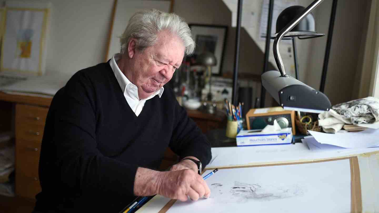 An old white man with a white side parting is sitting at a drawing table and is erasing a pencil drawing