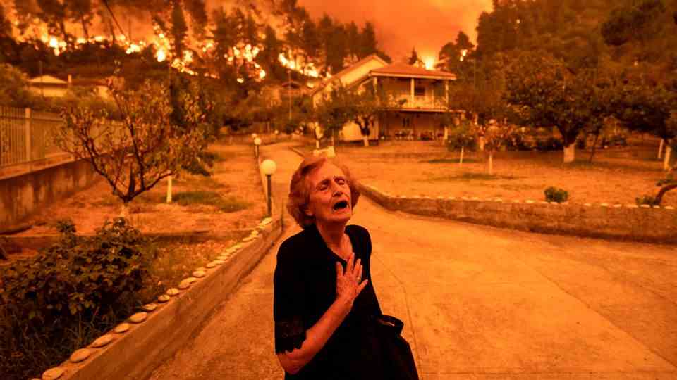 An old woman in black clutches her chest as a forest fire approaches houses in the background