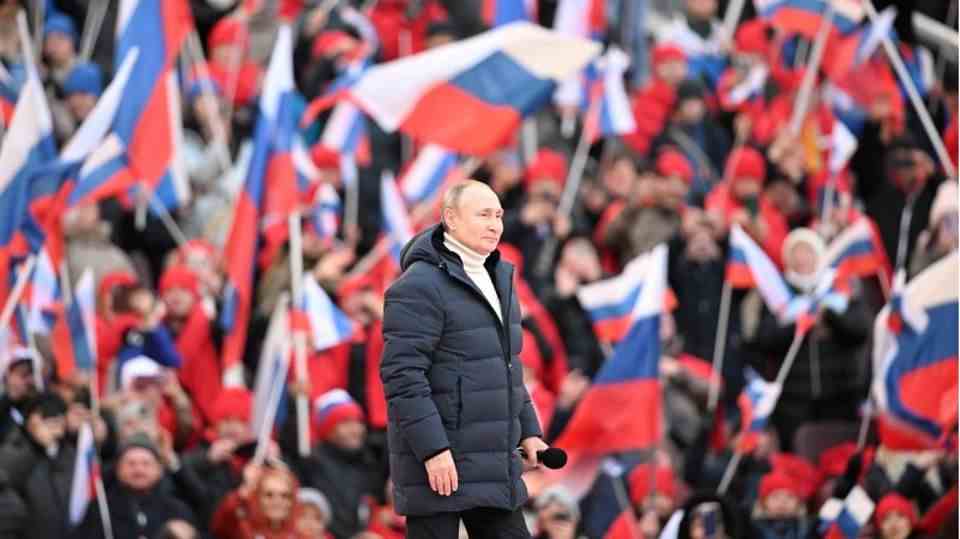 Vladimir Putin surrounded by people with Russian flags, wearing a lined jacket