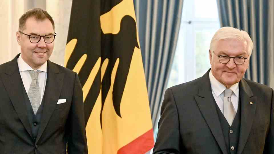 Two white men in tails stand in front of a flag: a black federal eagle on a yellow background with a thin red border