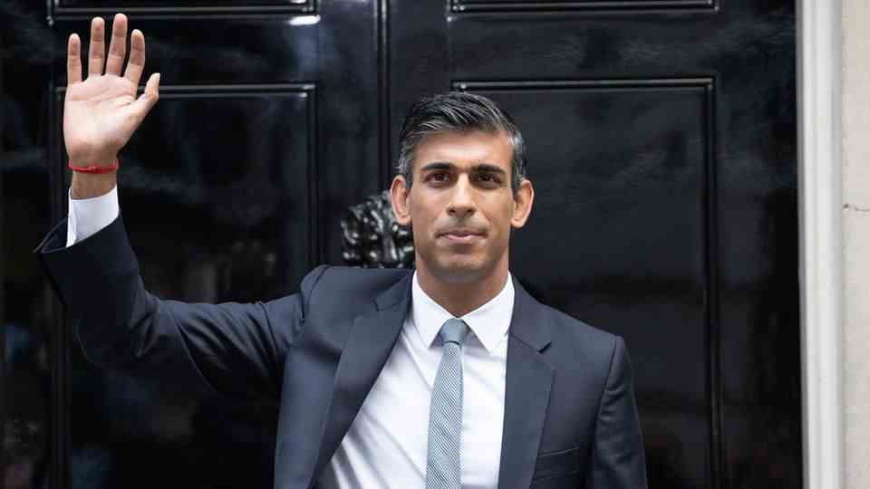 A man of Indian origin is standing in a suit and tie in front of a black lacquered front door with a 10