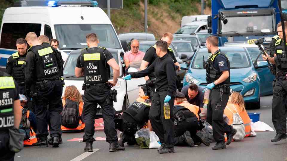 Young people in orange safety vests sit between cars on a multi-lane road.  Policemen stand behind them