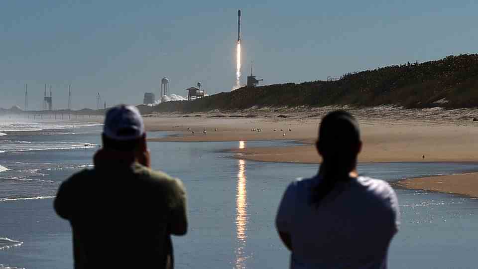 Two men watch as a SpaceX Falcon 9 rocket launches from Pad 39A at Kennedy Space Center from Canaveral National Seashore.  The rocket carries 49 Starlink internet satellites for a broadband network.