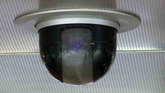 People in a train station are reflected in the lens of a surveillance camera.  © NDR 