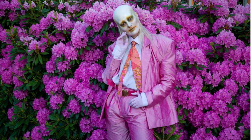 Singer Karin Dreijer from Fever Ray in a pink suit, with white make-up on her face, in front of a bush with pink flowers