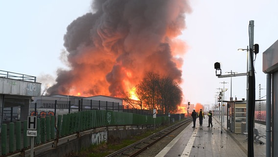 Police officers stand at a bus stop at Hamburg-Rothenburgsort train station while flames from a major fire can be seen in the background.  © Jonas Walzberg/dpa 