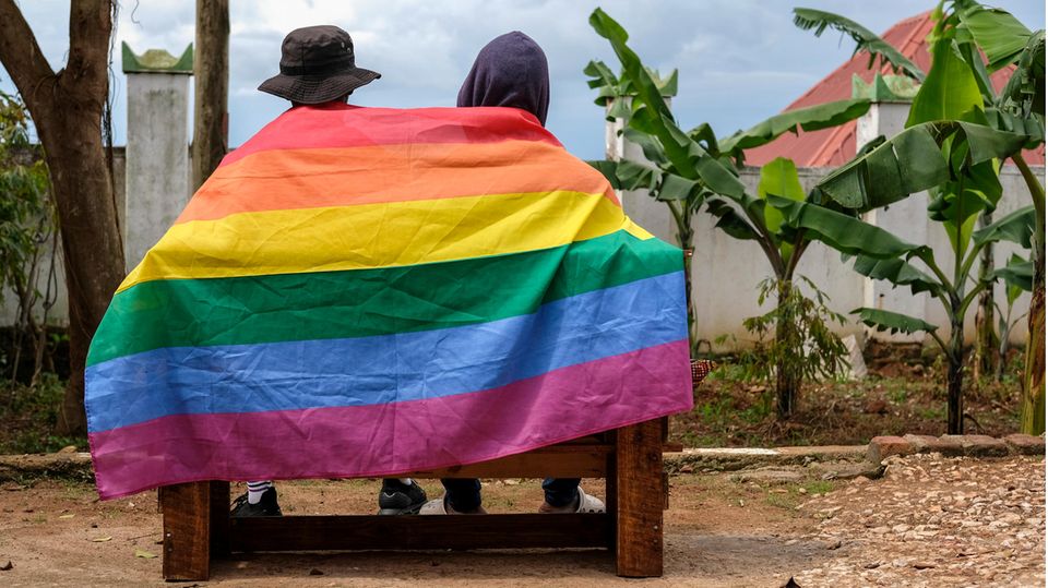 Two people in Uganda are seen from behind on a bench, their backs covered by a rainbow flag