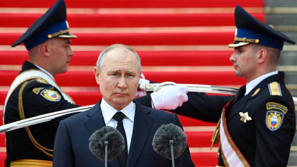 Putin stands in front of a microphone, behind him two soldiers
