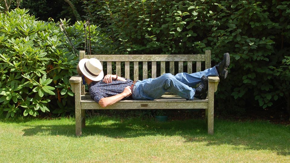 A man is resting on a bench.  He protects his face from the sun with a hat.