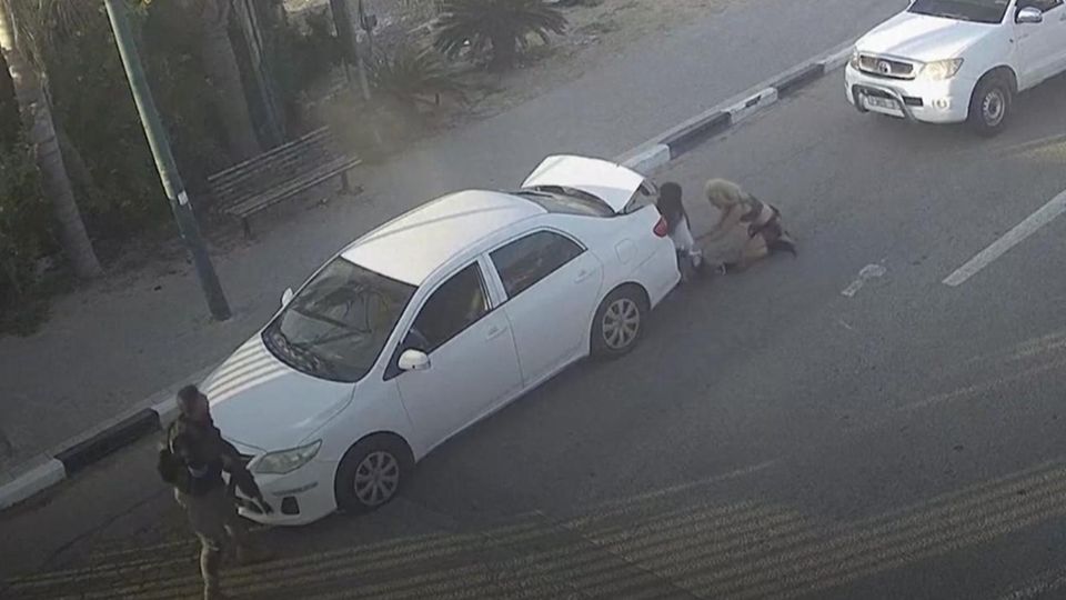 Two women hide behind a car, an Israeli police officer on the left