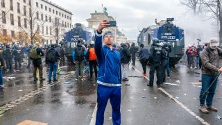 A man takes a selfie in front of a demonstration in Berlin, with water cannons in the background (Source: dpa/Vladimir Menck/SULUPRESS.DE)