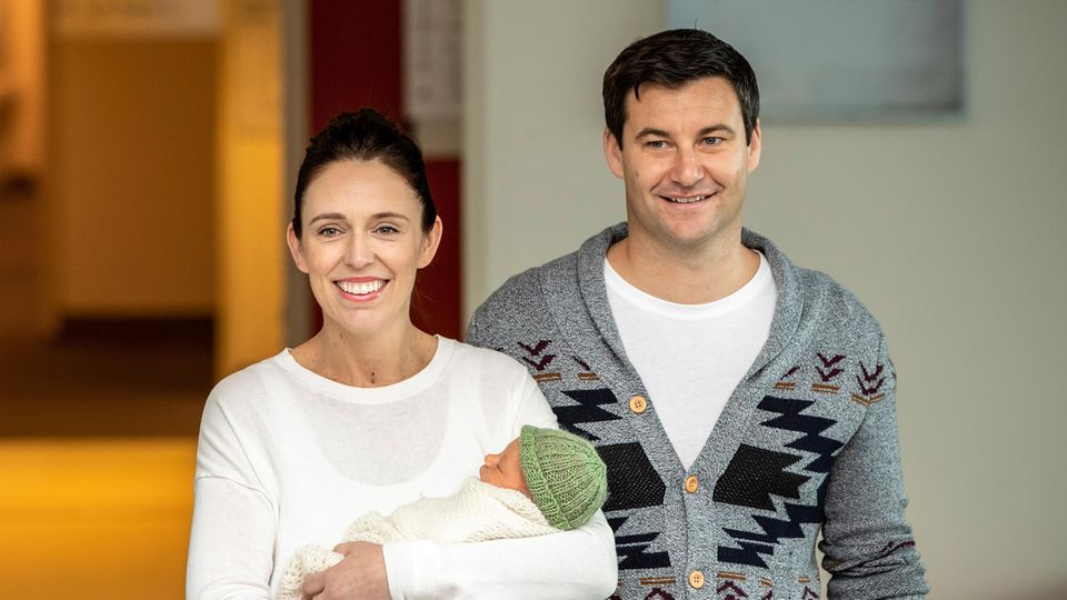Jacinda Ardern stands in front of a house with a baby in her arms.  Her partner Clarke Gayford stands next to her