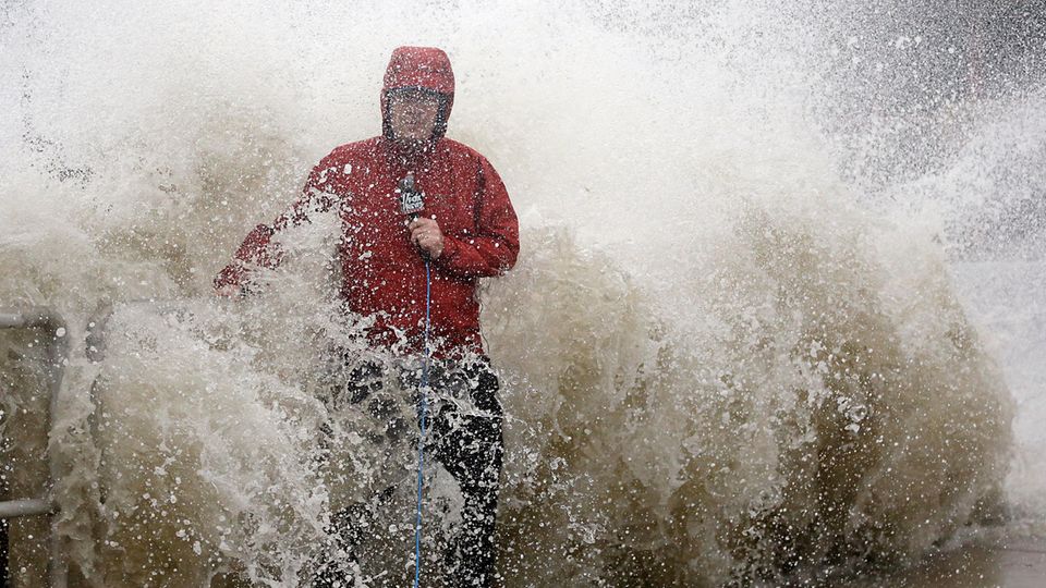 A reporter in a red jacket is surprised by a sideways wave