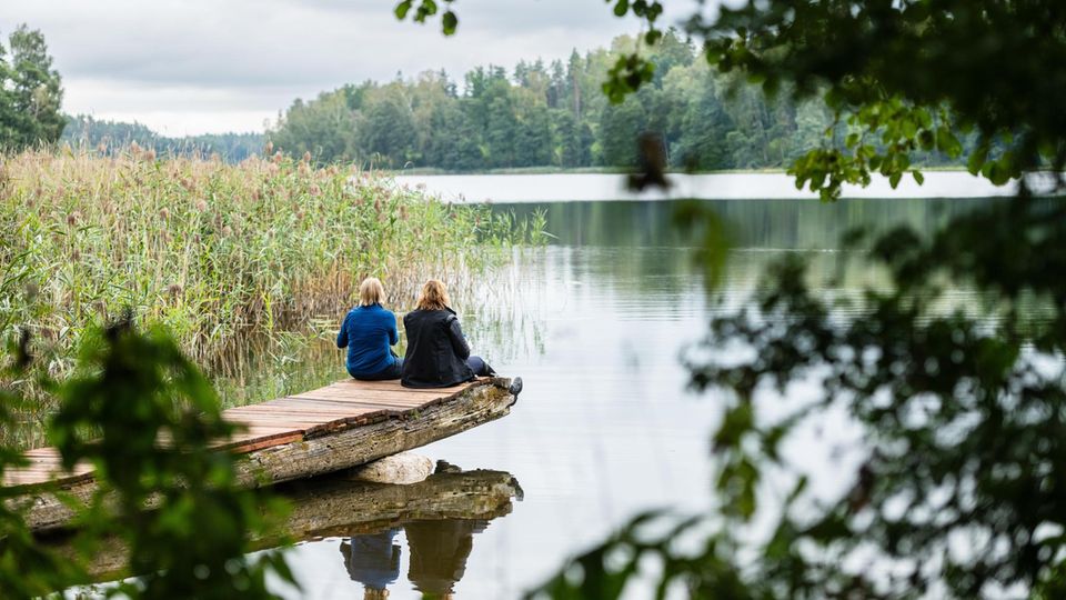 zwei Frauen mit dem Rücken zur Kamera sitzen auf einem Steg an einem See
