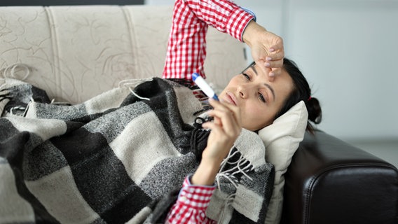A sick woman lies on a sofa with a wool blanket and looks at a thermometer.  © Colourbox 