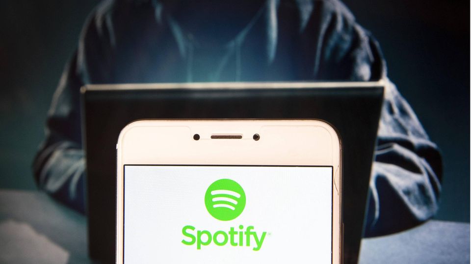 The Spotify logo on a smartphone screen with a person wearing a hood sitting behind it