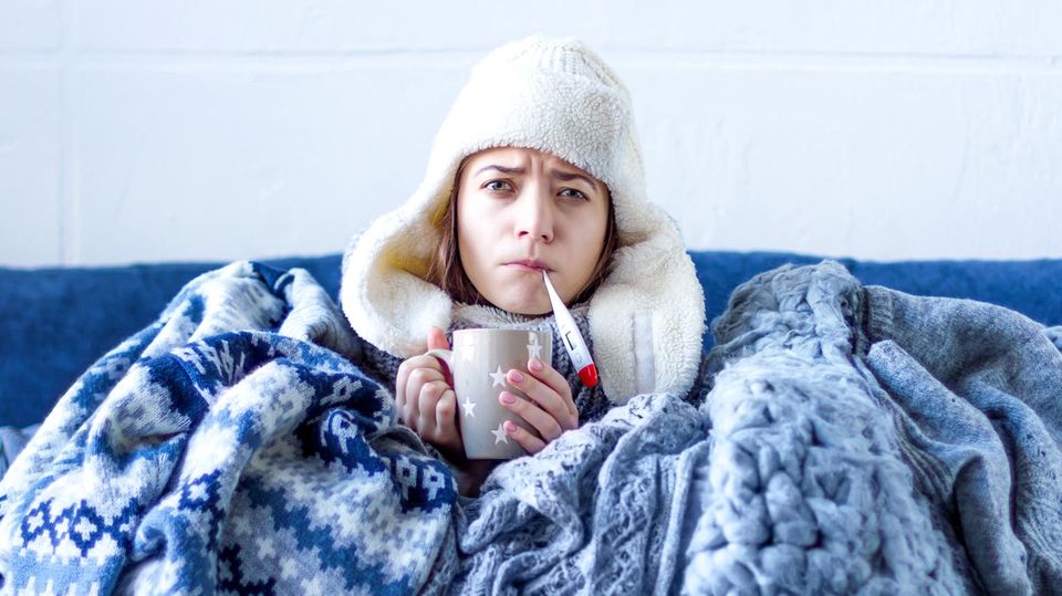 Woman with a cold sits on the sofa with a wool blanket and hat