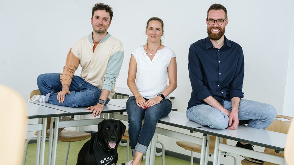 Three young teachers sit on a desk with the school dog, a black Labrador, at their feet