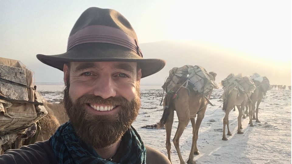 A man with a hat and a dark beard looks into the camera and smiles, behind him are packed camels