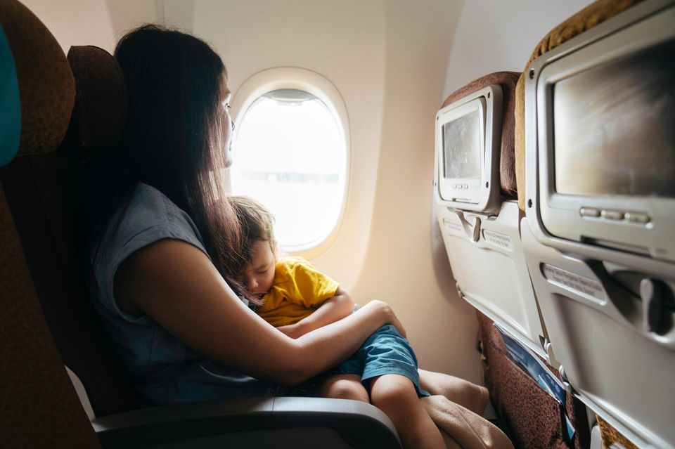 A woman sits on an airplane with her sleeping child on her lap and looks out the window.