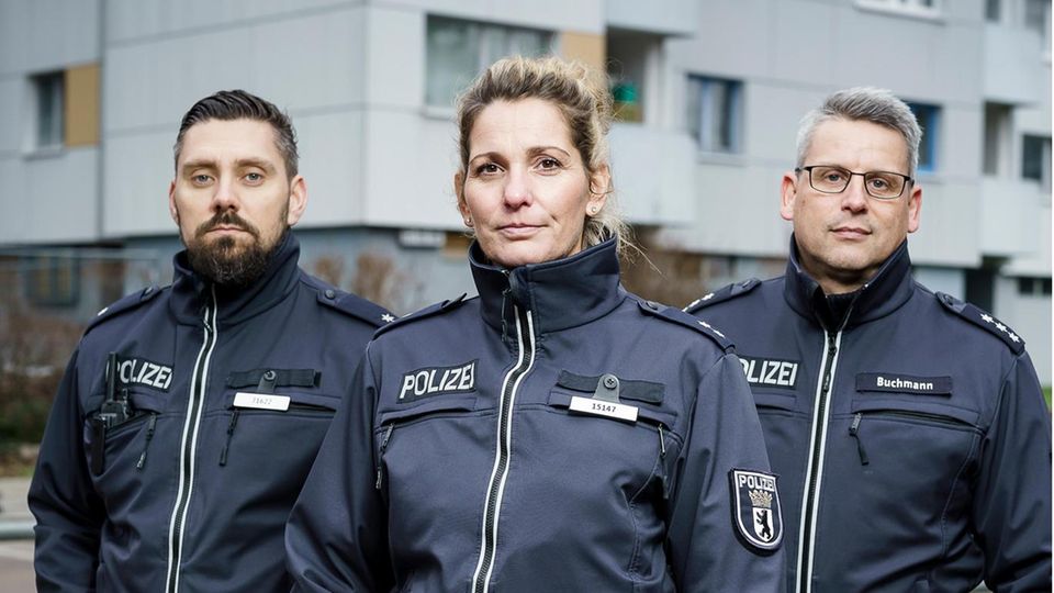 Three Berlin police officers (two men and one woman) stand in front of a high-rise building in Neukölln