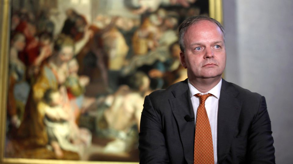 Eike Schmidt stands seriously in front of a painting that can be seen blurry in the background