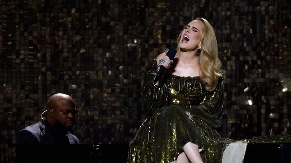 Adele sits on a grand piano at a concert.