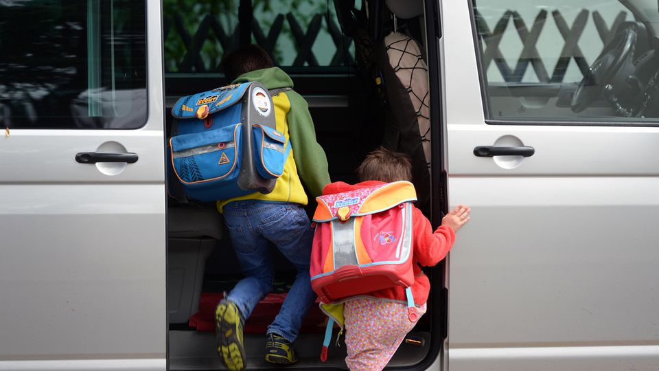 Two elementary school children with satchels on their backs climb into the sliding door of a silver VW bus
