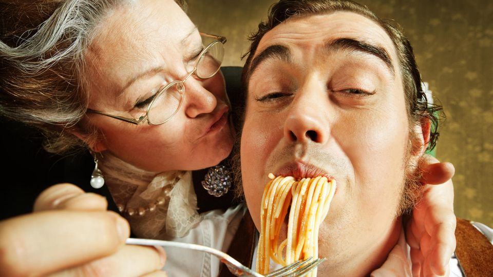 An Italian mom kisses her adult son on the cheek while feeding him spaghetti